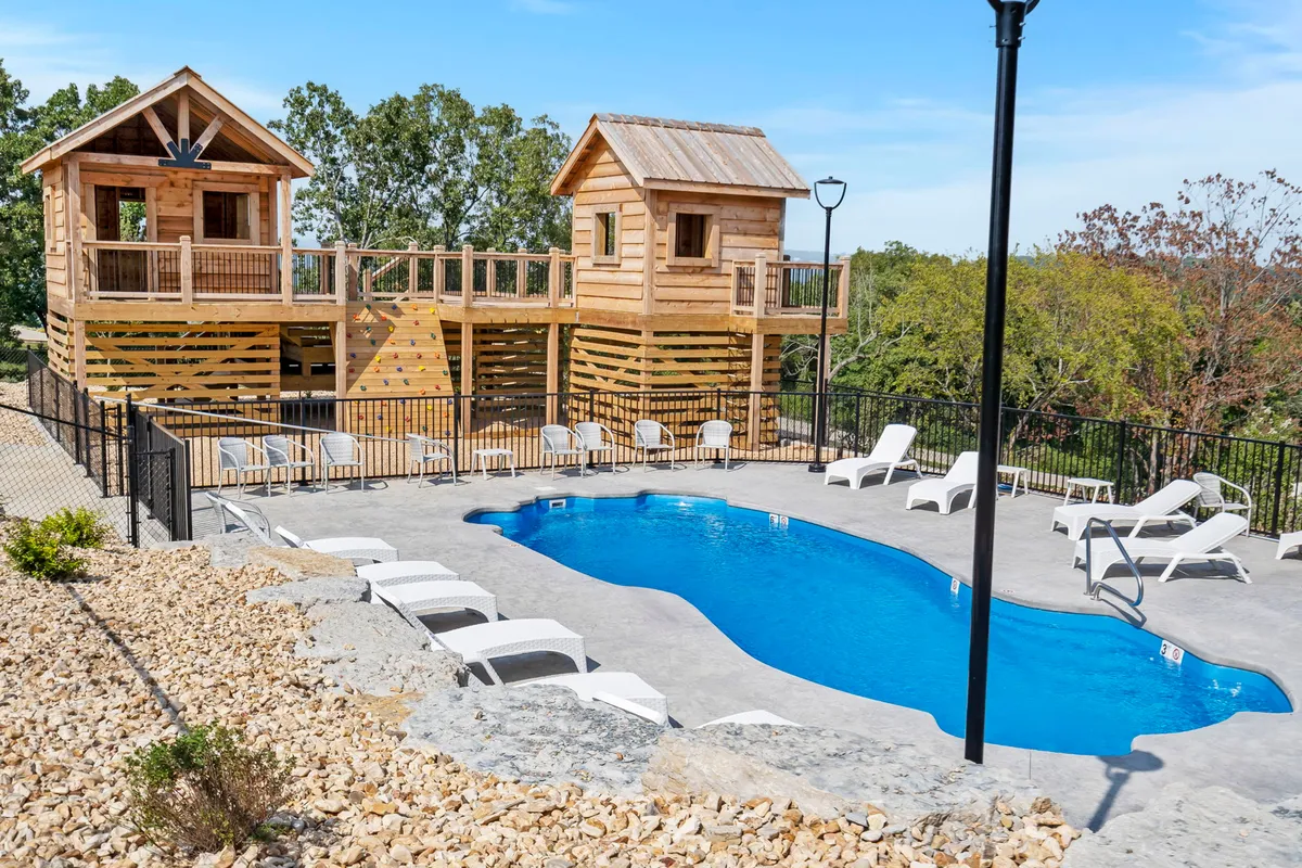 An outdoor setting showing a swimming pool surrounded by lounge chairs, two wooden playhouses, and lush greenery. The mood is lively and relaxed, with spaces for both active play and quiet relaxation under the sun.