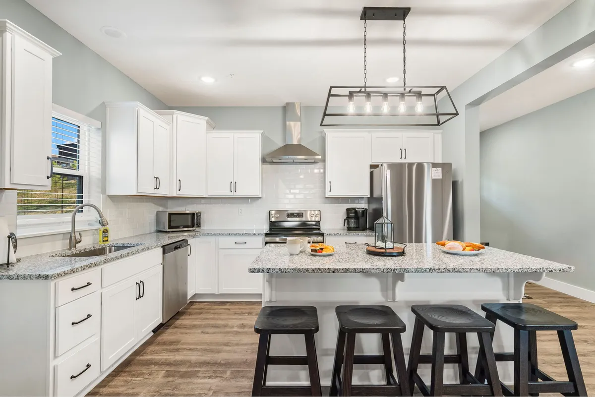 A bright, contemporary kitchen with a granite island, stainless steel appliances, white cabinetry, and modern light fixture. Stools at the island invite casual breakfasts or group cooking, and the kitchen exudes functionality and warmth.