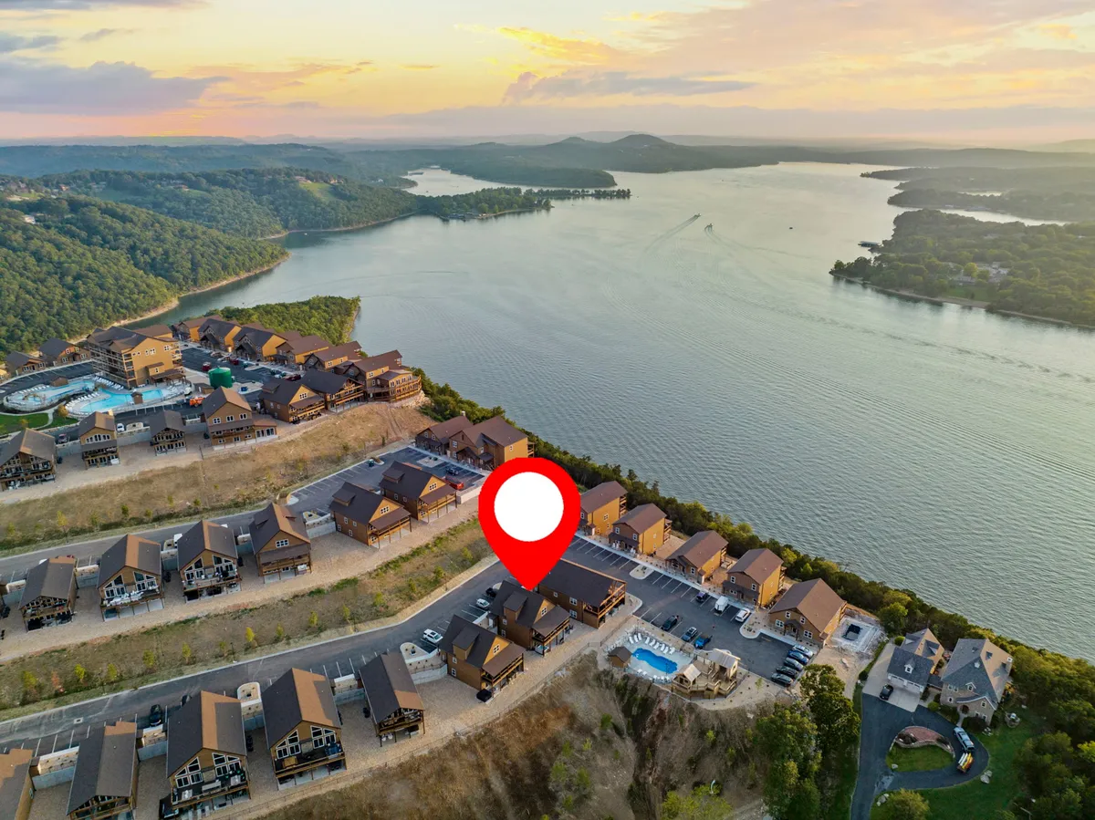 A scenic exterior view of Table Rock Lake surrounded by rolling Ozark hills at sunset, with residential buildings and a swimming pool in the foreground. The tranquil water reflects the colorful sky, and the natural landscape stretches out into the distance.