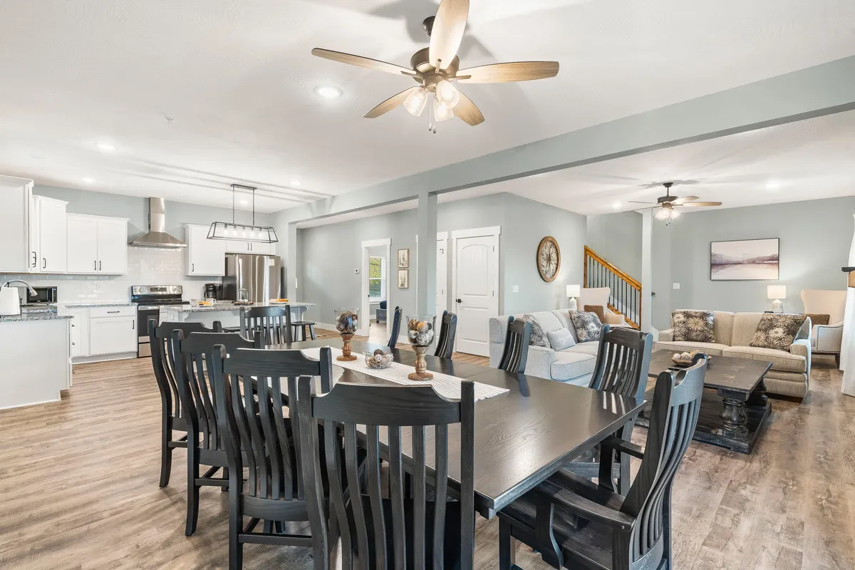 The interior scene centers on an inviting dining table, adjacent living room seating, and a kitchen island. The arrangement suggests a communal space designed for family dinners, entertaining guests, and casual relaxation, with the ceiling fan adding a touch of homey comfort.