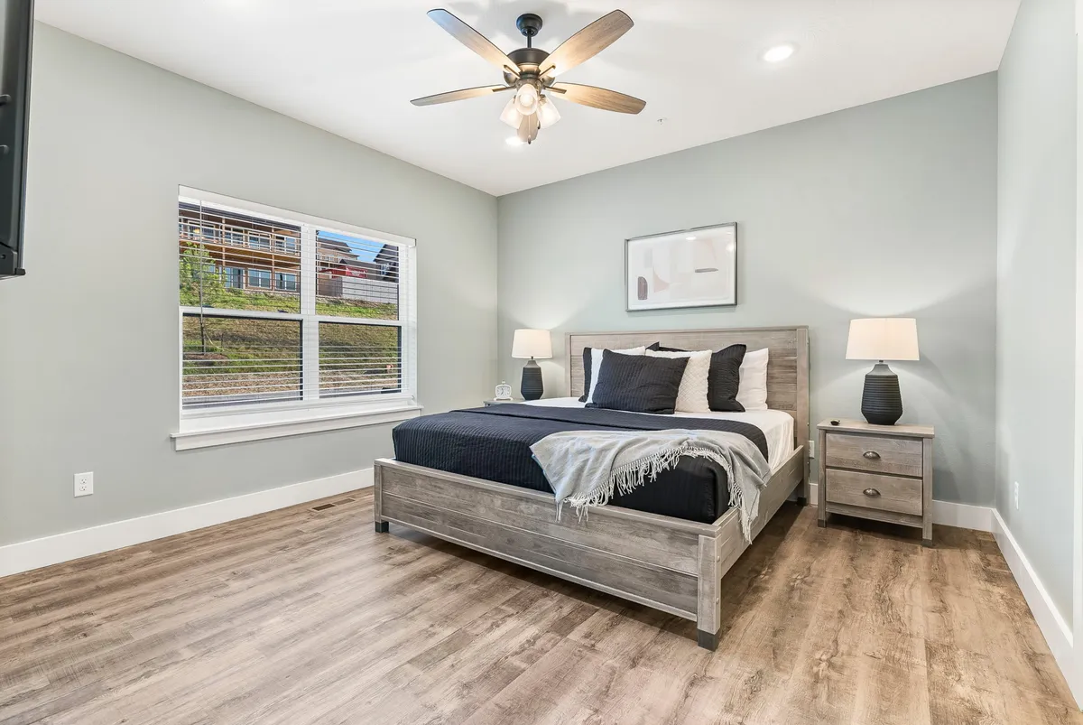 A tranquil bedroom featuring navy blue bedding, a window with blinds allowing natural light to filter in, a nightstand with a lamp, and tasteful wall art. The space is neat, airy, and exudes a sense of calm and comfort.