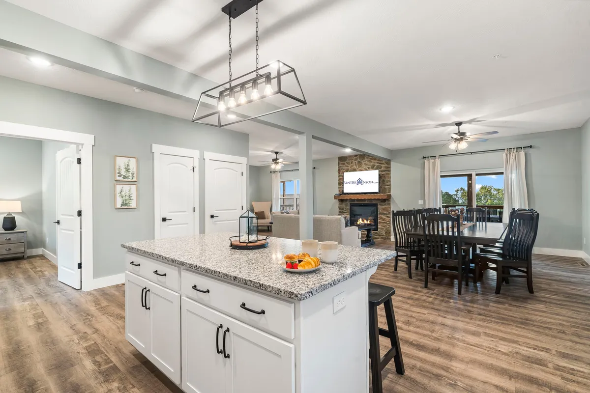 This image presents the heart of the home with a clear view of the open-concept space encompassing the granite kitchen island, dining table, stone fireplace, and large windows with scenic lake views. Warm lighting and inviting décor evoke a communal, welcoming feel.