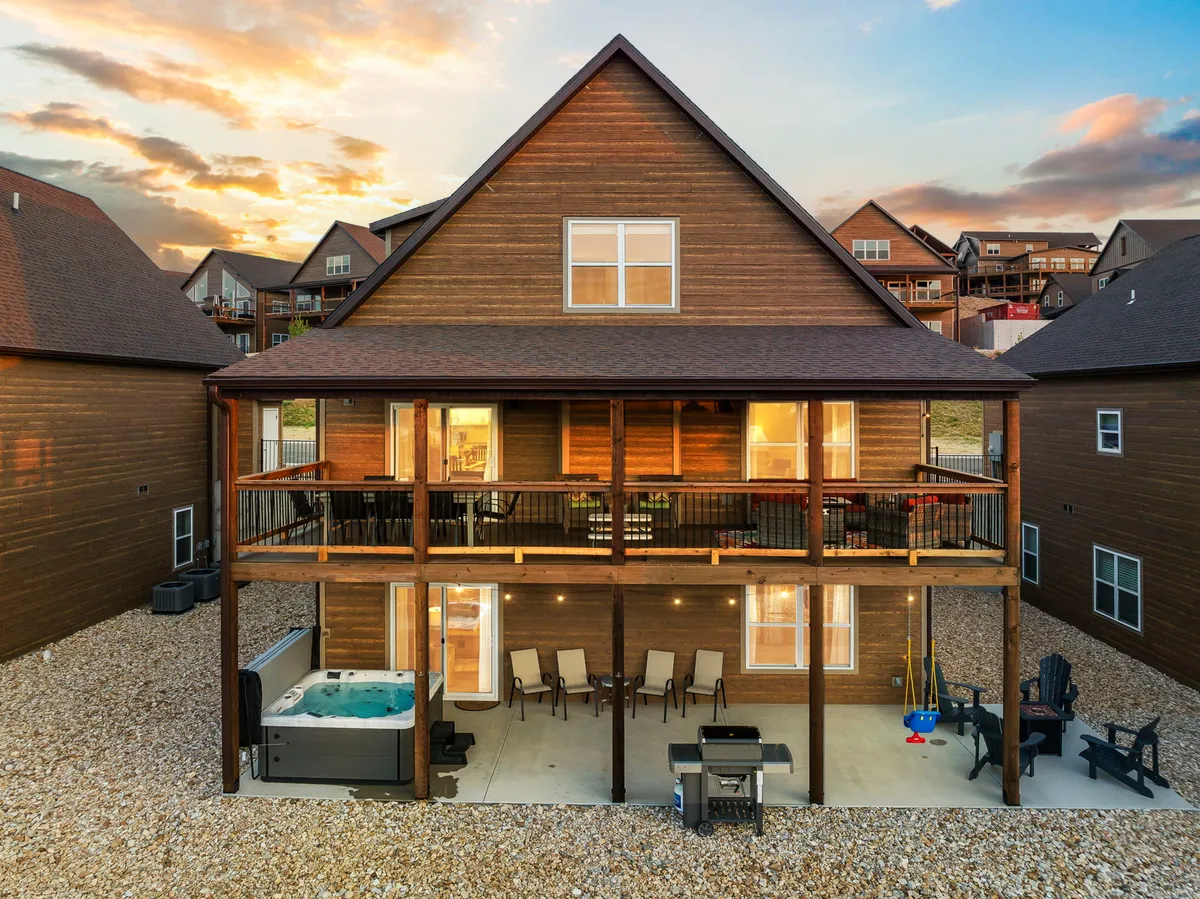 The image showcases the inviting outdoor living space of a spacious single-family house perched atop Table Rock Lake. The focal points include a bubbling private hot tub with scenic lake views, a cozy outdoor seating area perfect for gatherings, a barbecue grill ready for alfresco feasts, and a swing set for kids. The warm, relaxed ambiance is heightened by the golden sunset glow across the deck, inviting guests to unwind and make memories together.