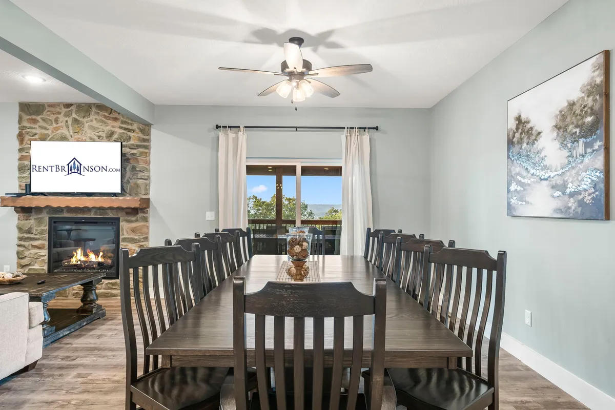 A spacious dining area centers around a large table, adjacent to a beautiful stone fireplace and flanked by expansive windows. The space flows naturally into the kitchen and living room, with cozy decor and abundant natural light.
