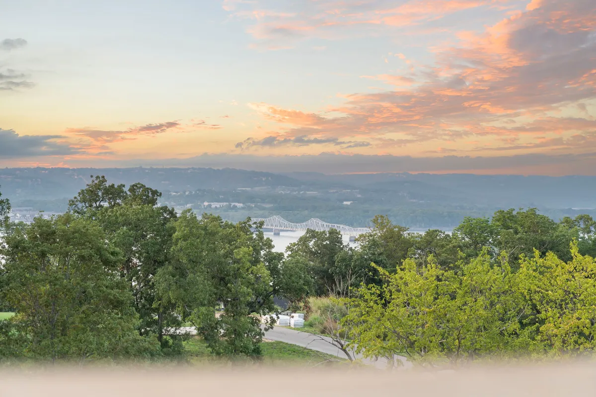 A serene natural view with a distant bridge spanning a peaceful river, framed by lush trees and a colorful sunset sky. The scene evokes a sense of calm, openness, and immersion in the Ozarks’ natural beauty.