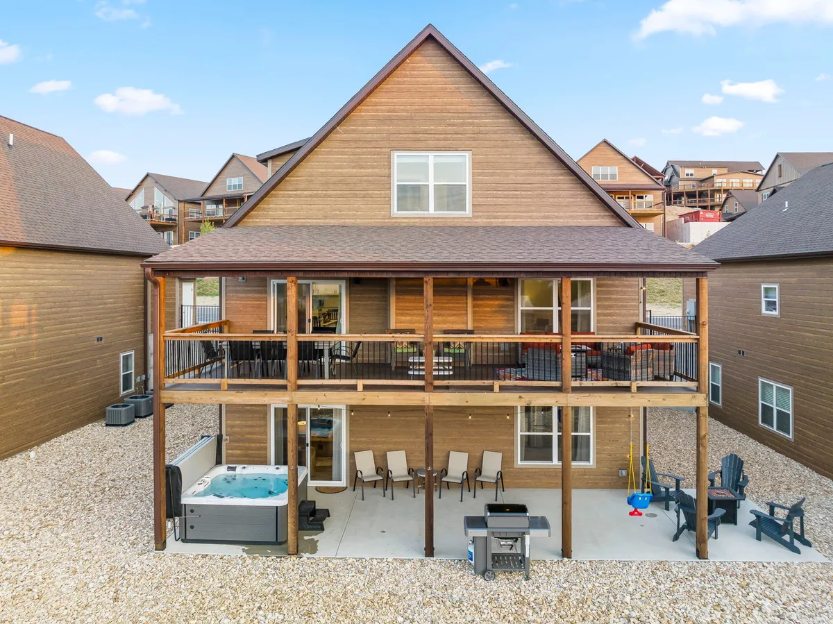 This image features a two-story wooden house with a hot tub, outdoor seating area, and a grill. The spacious patio invites guests to relax, dine, or socialize while enjoying sunset views from the balcony and the ambiance of an outdoor gathering space.